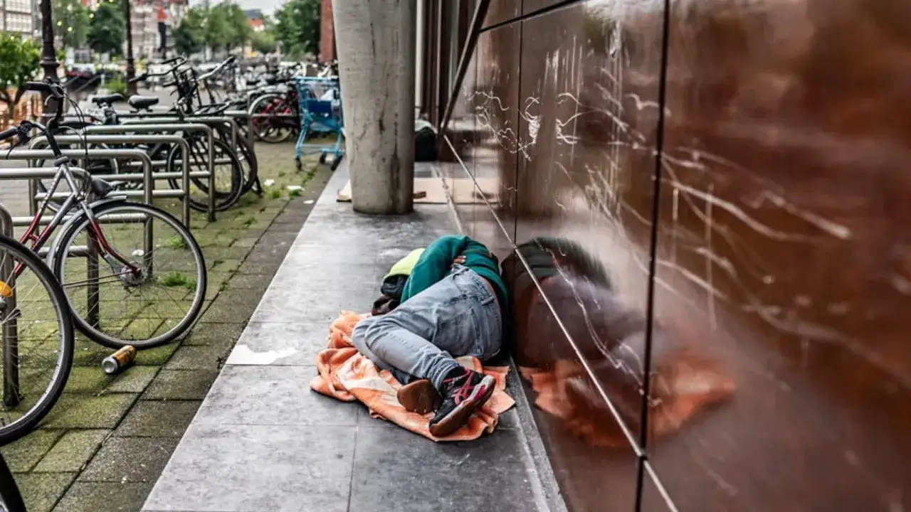 Een persoon slaapt op een deken naast een gebouw, omringd door fietsen. Een schrijnend beeld van daklozen in Nederland.