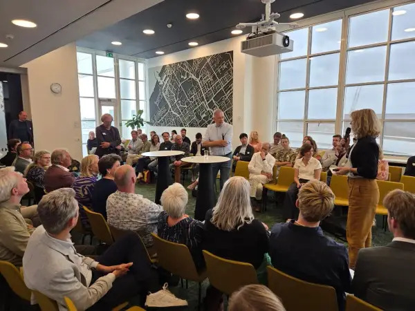 Mannen en vrouwen luisteren aandachtig naar een spreker, mogelijk over ruimtelijke ordening, in een zaal met een grote stadskaart aan de muur.