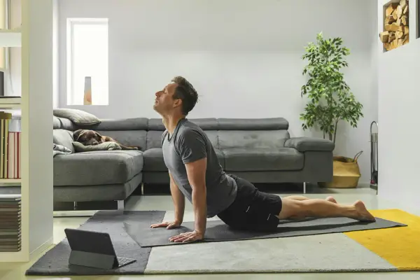 Man in cobra pose op yogamat, neemt regie over zijn leven. Hond slaapt op bank.