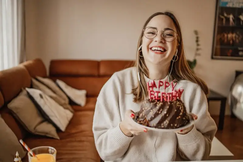Vrolijke vrouw met bril en koptelefoon viert haar verjaardag met een chocoladetaart. Ze heeft de volwassen leeftijd bereikt en straalt van geluk.